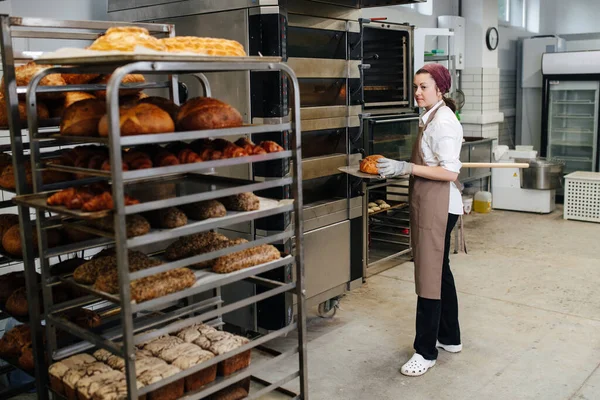 Düz bir kürek üzerinde taze ekmeği olan fırıncı bir kadın. Saç bandı takıyor. Fırın mutfağının içinde..