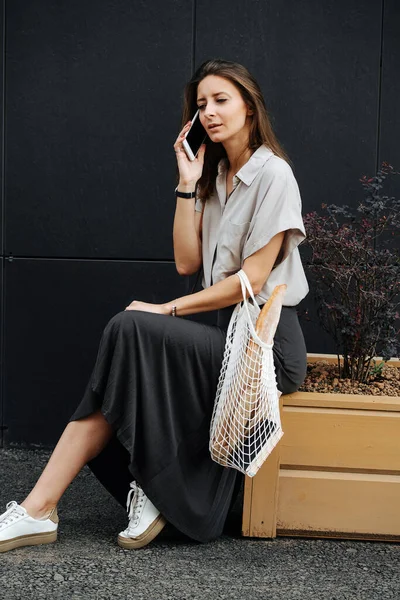 Mature woman in corporate clothing on the street. She\'s talking on the phone, sitting on a planter box with bonsai tree, holding net bag with baguette.