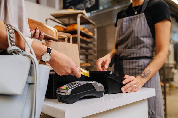 Woman making a purchaise, paying with her credit card. Baker standing behind cash register calculating price. Cropped, no heads.