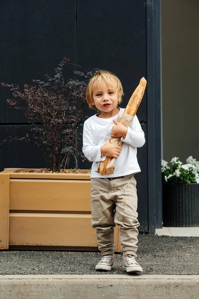 Little boy with blond hair holding baguette with both hands, outdoors on the street. Over back panels, next to planter box.