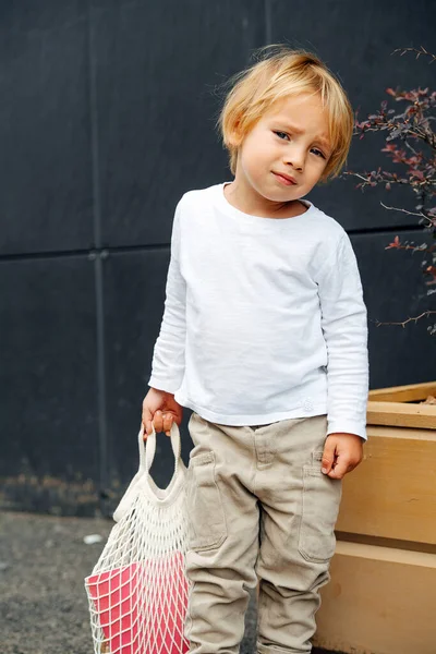 Little boy with blond hair holding net bag with toy bucket, outdoors on the street. Over back panels, next to planter box.
