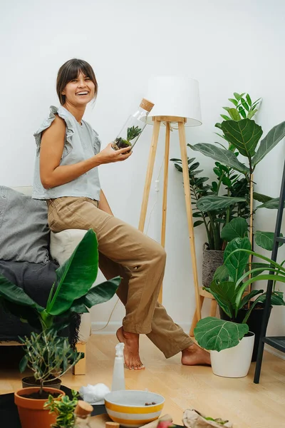 Carefree social woman sitting on a sofa, holding glass bottle with small plant — Stock Photo, Image