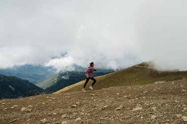 Dağların tepelerinde tepelere doğru koşan, kısa çimenler ve ağaçlarla kaplı, alçak bulutlarla çevrili genç bir kadın. Kapüşonlu, yoga pantolonlu ve sırt çantalı. Yan görünüm.