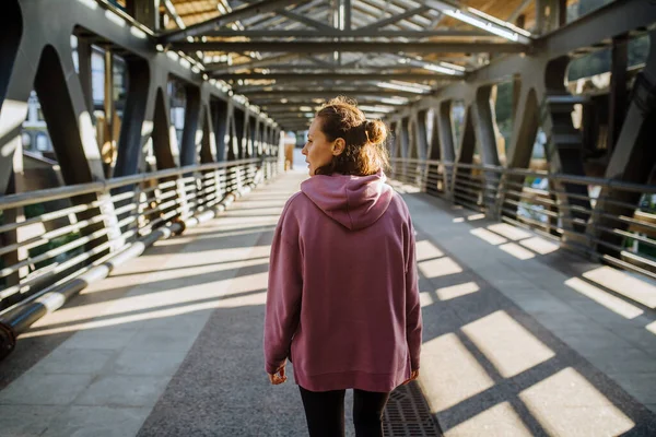 若い女性は屋根と鋼の桁の橋を歩いて渡る ピンクのパーカーとヨガパンツを着てる 後ろから — ストック写真