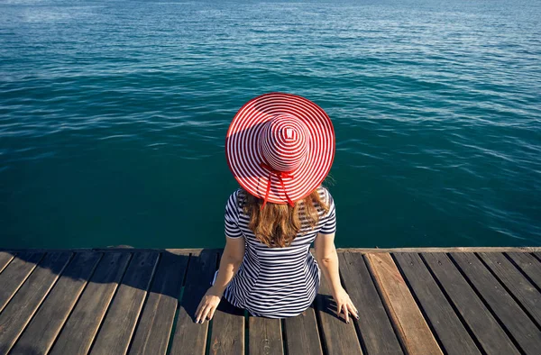 妇女在帽子和条纹礼服坐在码头上 看着翡翠蓝海 夏天和海概念 — 图库照片
