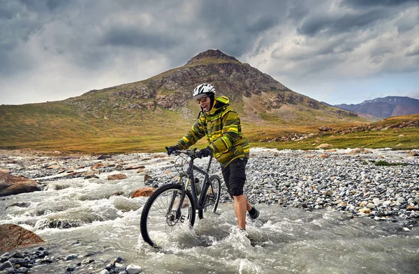 Man with mountain bike crossing the river in the wild mountains against cloudy sky background
