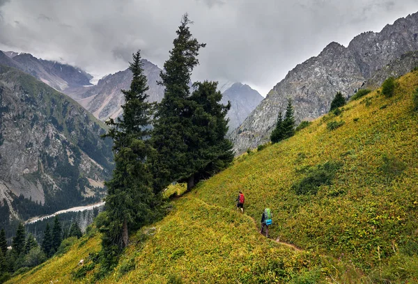 Two tourists with backpacks walking on the trail in the mountain valley at overcast sky background. Travel adventure concept