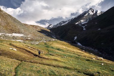 Sırt çantalı küçük turist, karlı zirveleri ve bulutlu arka planı olan yeşil dağ vadisinde yürüyor.