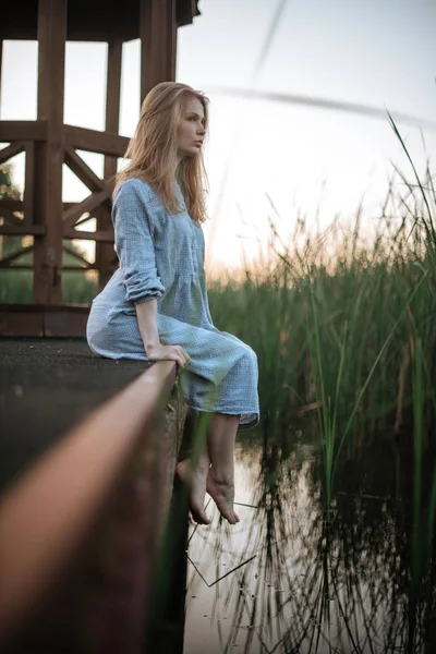 Romantisch meisje in blauwe jurk zitplaatsen op de pier. Vegetatieve achtergrond — Stockfoto