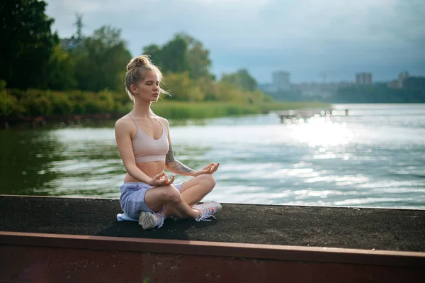 Cvičení jógy a meditace v přírodě při východu slunce. Žena cvičí poblíž města na molu. — Stock fotografie