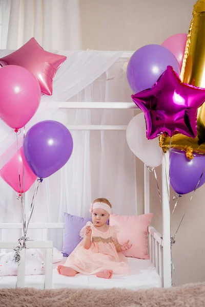 Bella bambina seduta in un letto in un vestito di compleanno mordere una bacchetta da un palloncino Fotografia Stock