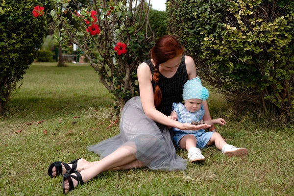 女の子は手のひらに子にバンプを示す 赤ちゃんに興味を持って見えます 赤い花を持つブッシュ大統領に対して 草の上の公園で座る ストック写真