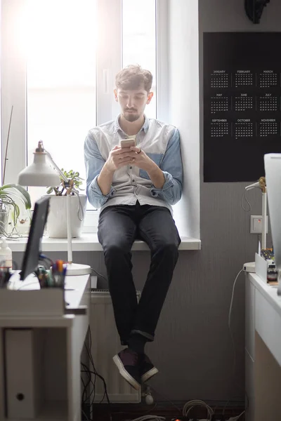 Joven hombre elegante sentado en la ventana con el teléfono en las manos — Foto de Stock