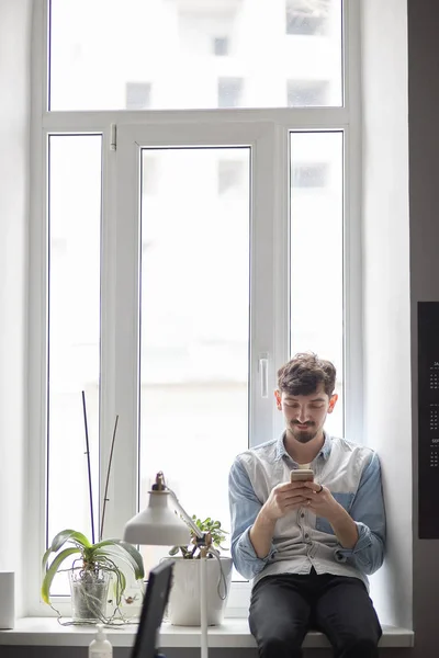 Giovane uomo elegante seduto sulla finestra con il telefono in mano — Foto Stock