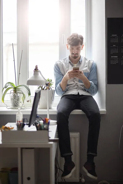 Stilvoller junger Mann sitzt mit Handy in der Hand am Fenster — Stockfoto