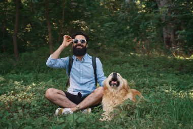 Neşeli adam oturuyor bacak bacak çimenlerin üzerinde onun güneş gözlüğü burnuna sabitleme, poz, köpeği ile birlikte kameraya bakıyor. Mutlu erkek ve hayvanını eğleniyor