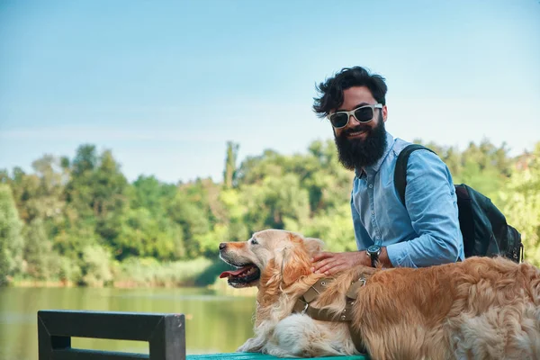 Best friends' concept - between human and animal. Side view, young man hugging, tapping and sitting with his dog on the chair in the park, enjoying the view of the lake, man looking to camera smiling.
