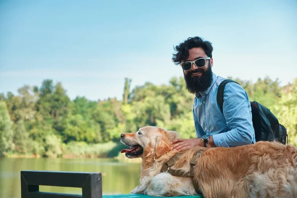 Arkadaşlarım kavramı - insan ve hayvan arasındaki. Yan görünüm, sarılma, dokunarak ve parkta, sandalyeye manzarayı Gölü, kamera gülümseyerek bakan adam köpeğiyle oturan genç adam.