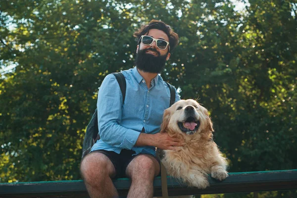 Köpeğiyle, altın geri almak, parkta, ağaçlar ve güneş ışığı arka planda sandalyeye oturmuş sakallı, şık yakışıklı hipster neşeli hayvanını masaj yaparken mutlu gülümseyerek. 