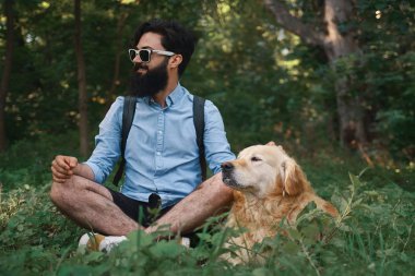 Yakışıklı adam çimlere oturup istirahat sevimli köpeği ile bacak bacak, gülümseyerek, her ikisi de doğru aynı yöne bakıyor, gün mükemmel. İnsan ve hayvan arasındaki dostluk. Mutlu arkadaş.