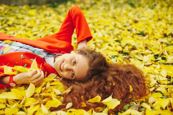 Happy girl laying in yellow autumn leaves with closed eyes.Smiling girl with long wavy hair enjoying life in autumn forest. Cheerful girl in red coat and colorful plaid shirt on beautiful fall day.