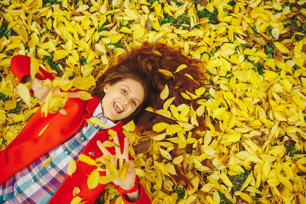 Autumn leaves falling on happy young woman in forest. Beautiful happy girl laying in yellow autumn leaves.