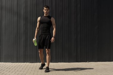 Elinde bir su şişesi ile gri metalik duvara poz atletik modern genç adam. Hidrasyon ve beslenme 's kavramı. Fitness kaslı erkek. İnsan dağ güç pozlar.