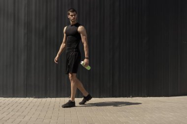 Elinde bir su şişesi ile gri metalik duvara poz atletik modern genç adam. Hidrasyon ve beslenme 's kavramı. Fitness kaslı erkek. İnsan dağ güç pozlar.