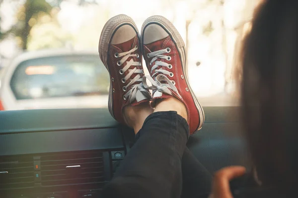 Modern woman tourist, photographer relaxing in the car with legs in sneakers on the glove compartment, holding a camera in hands looking to screen.