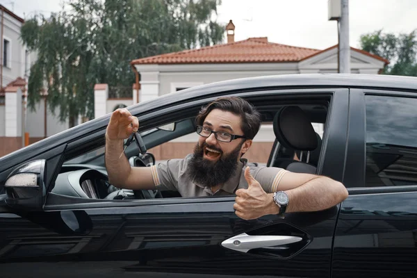 Car dealership, the happiest client. Charismatic man holding car keys showing thumbs up and smiling, with teeth, while sitting in the car looking to camera through the window..
