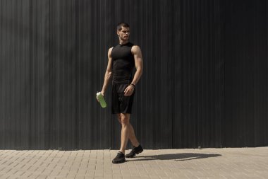 Elinde bir su şişesi ile gri metalik duvara poz atletik modern genç adam. Hidrasyon ve beslenme 's kavramı. Fitness kaslı erkek. İnsan dağ güç pozlar.