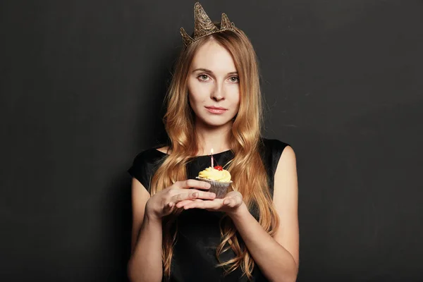 Pretty young woman, with a crown, holding a birthday cupcake with a candle  and cherry on top on black background