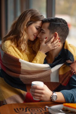 Kucaklayan ve öpüşme genç çift sıcak bir ekose battaniye ve bir fincan kahve arka planda windows ile kaplı