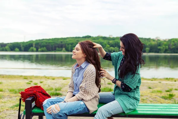 两个美丽的女朋友有乐趣 放松和微笑 褐发女郎做发型发红 而坐在长凳上与真棒绿色景观的背景 — 图库照片