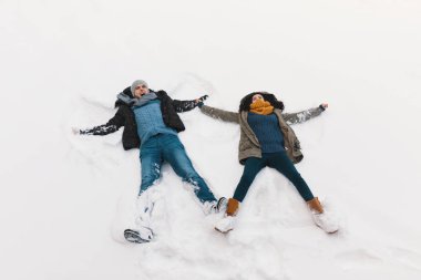 Carefree woman and man having fun while lying on the white snow in the position of flying angels outdoors during winter clipart