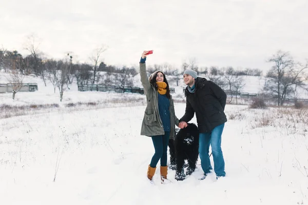现代无忧无虑的男人和女人在与他们蓬松的狗在冬季景观背景上牵手的同时 与他们的毛茸茸的狗做自拍 — 图库照片