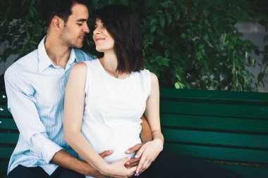 Genç modern bir çift arka planda o hamile ve yuvarlak karnı yavaşça beşik yaprakları ile bir bankta oturur. Onu yavaşça kucaklayan.