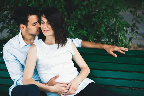 A young modern couple sits on a bench with leaves in the background she is pregnant and cradles her rounded belly gently. He embraces her gently.
