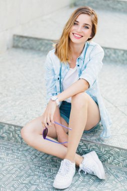 Işık gri basamaklarında oturan bir mutlu genç beyaz bir kadın portresi. Güzel gülüş, dudak, diş, mavi, güzel, gözler, kaşlar. Pozitif kişi kavramı