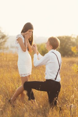 Onların hayatında genç benzersiz an ayakta ona teklif onun diz, dostum. Mutlu, gözleri, yüz altın alan arka plan güneş ışığı ile üzerinde el ile kapsayan inanamıyorum.