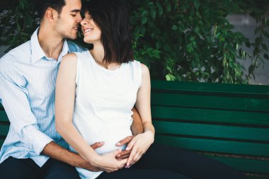 Genç modern bir çift arka planda o hamile ve yuvarlak karnı yavaşça beşik yaprakları ile bir bankta oturur. Onu yavaşça kucaklayan.