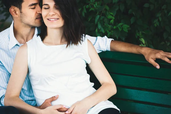 現代の若い夫婦は 彼女は妊娠している 彼女の丸みを帯びた腹を優しくあやし 背景の葉のベンチに座っています 彼は彼女を優しく包み — ストック写真