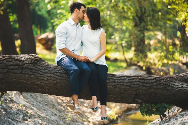 美丽的怀孕夫妇在森林外面放松 坐在溪边的树茎上接吻 背景是阳光穿透绿叶 — 图库照片