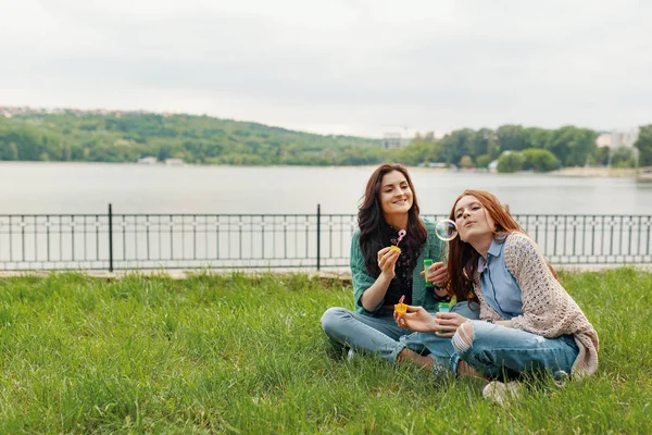 两个姐妹一边泡泡一边玩 坐在湖边的草地上 背景是美丽的风景 — 图库照片