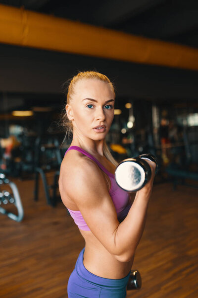 Young woman in pink top and mini shorts doing exercise with dumbbells while training arms at the gym 