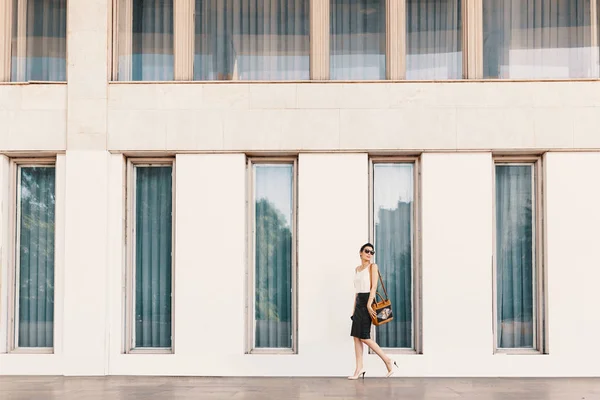 Модная высокая бизнес-леди в солнцезащитных очках прогуливается возле здания с высокими окнами в городе — стоковое фото