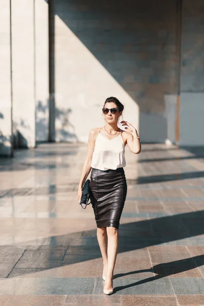 Refined young woman in leather skirt and silk blouse walking confident near a building.