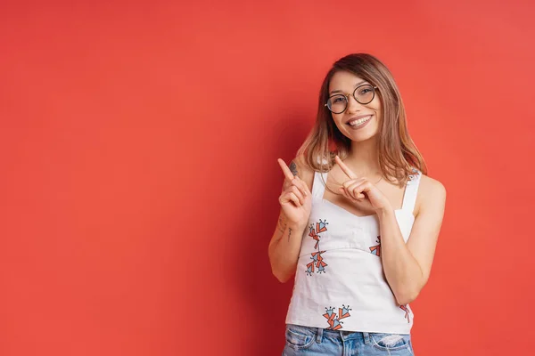 Mujer joven sonriente con gafas, señalando a un lado el fondo rojo — Foto de Stock