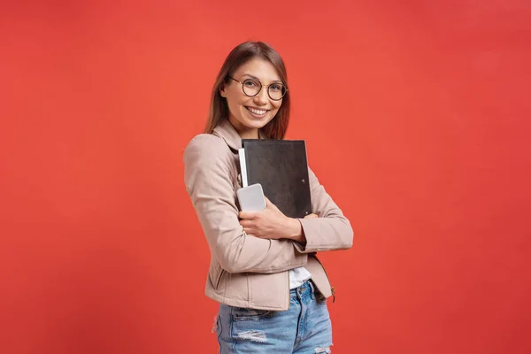 Νέος χαμογελαστός φοιτητής ή ειδικευόμενος σε γυαλιά που στέκονται με ένα φάκελο σε κόκκινο φόντο. — Φωτογραφία Αρχείου