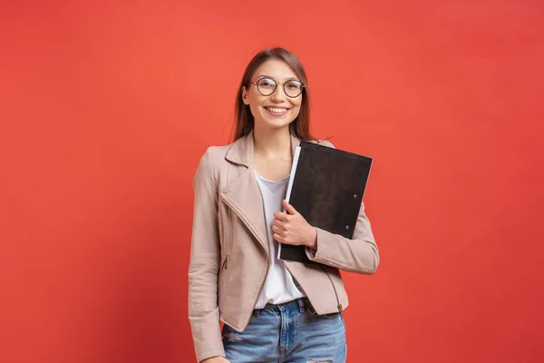 Ung leende elev eller praktikant i glasögon stående med en mapp på röd bakgrund. — Stockfoto